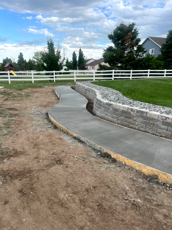 A freshly poured concrete pathway curves through a yard, bordered by a retaining wall and dirt.