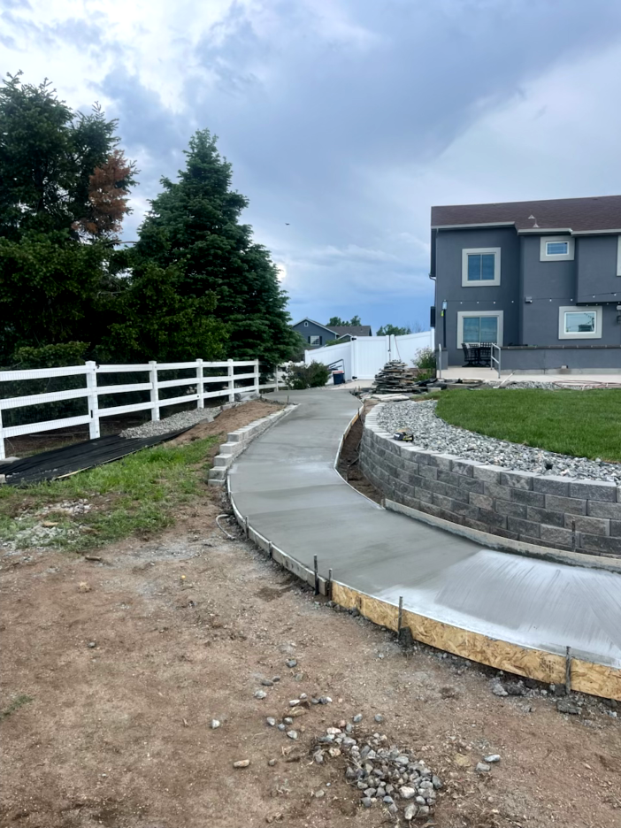 Newly poured concrete walkway curves toward a house. Wood forms, retaining wall and white fence are visible. Cloudy sky.