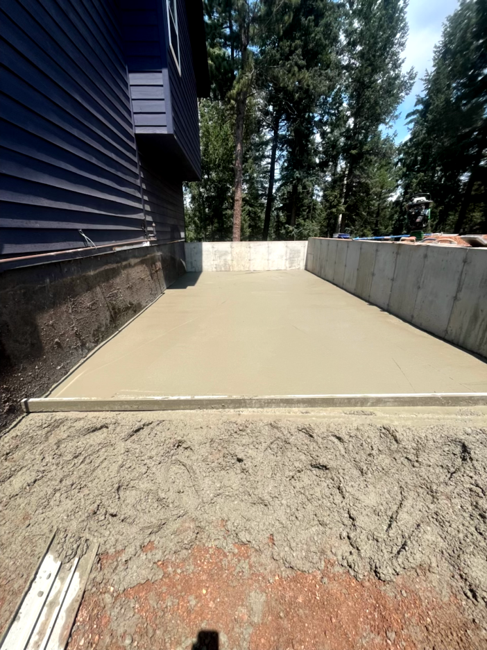Newly poured concrete patio next to a blue house and concrete retaining wall; dirt in foreground.