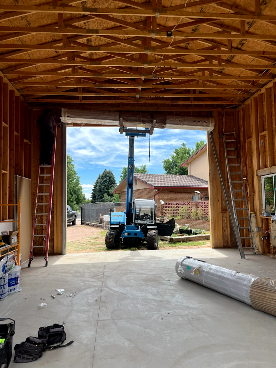 Garage under construction; blue lift inside doorway, framing exposed. Bright sky and backyard visible.
