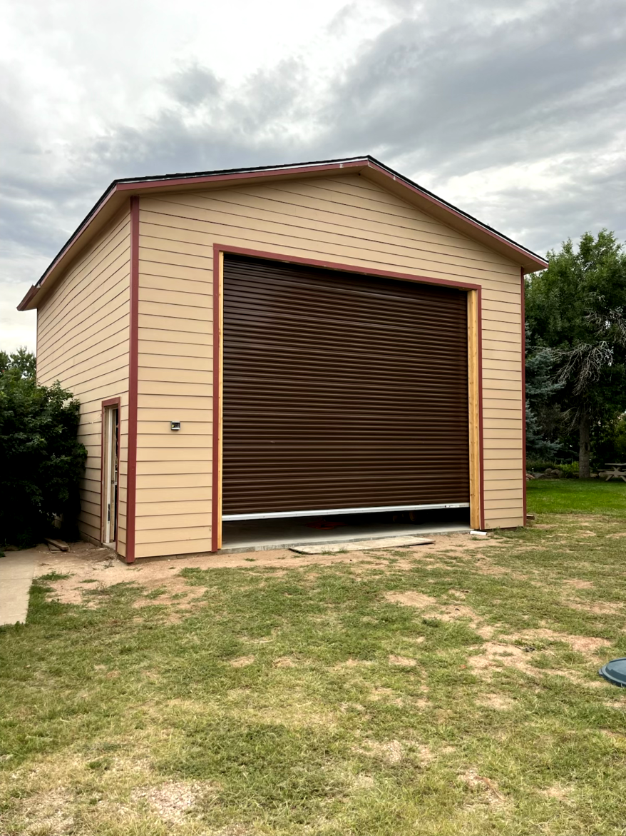 Brown roll-up garage door on tan building with red trim, in a grassy yard under a cloudy sky.