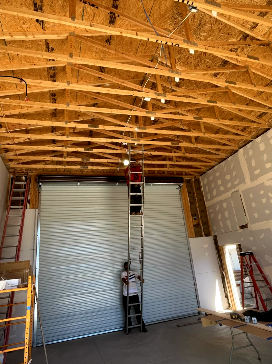 Interior of a building under construction. Person on a tall ladder near a roll-up garage door; unfinished ceiling.