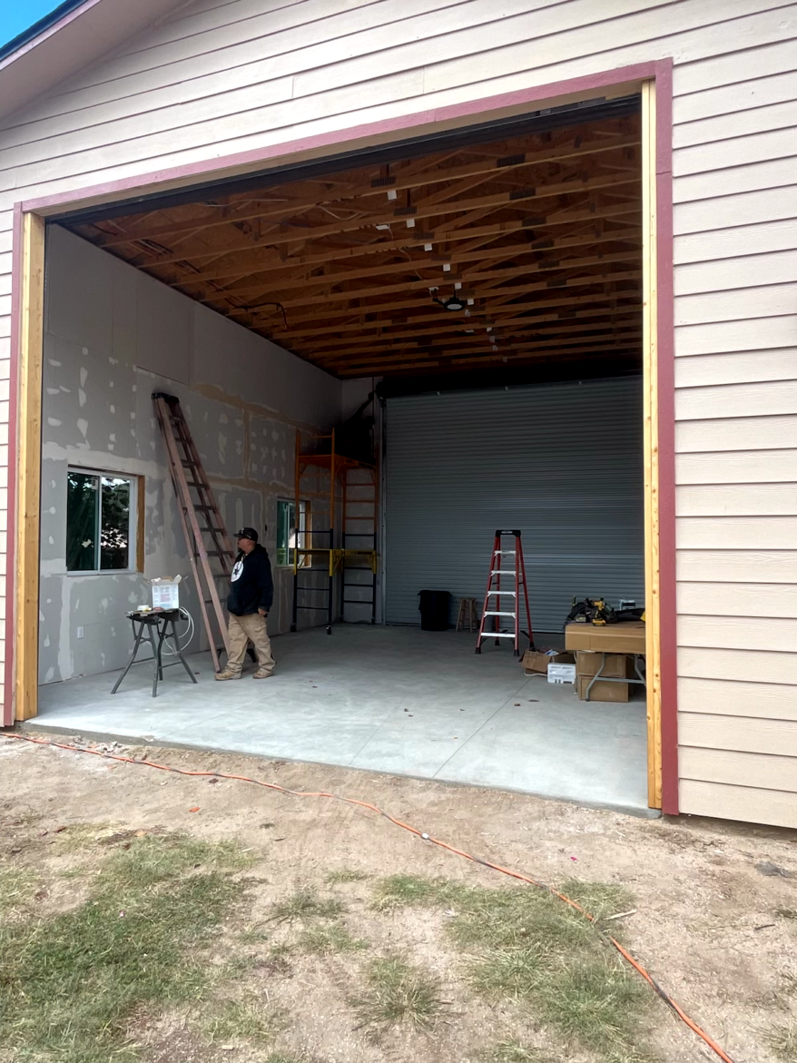 Open garage with person inside; unfinished interior, concrete floor, ladder, and equipment.