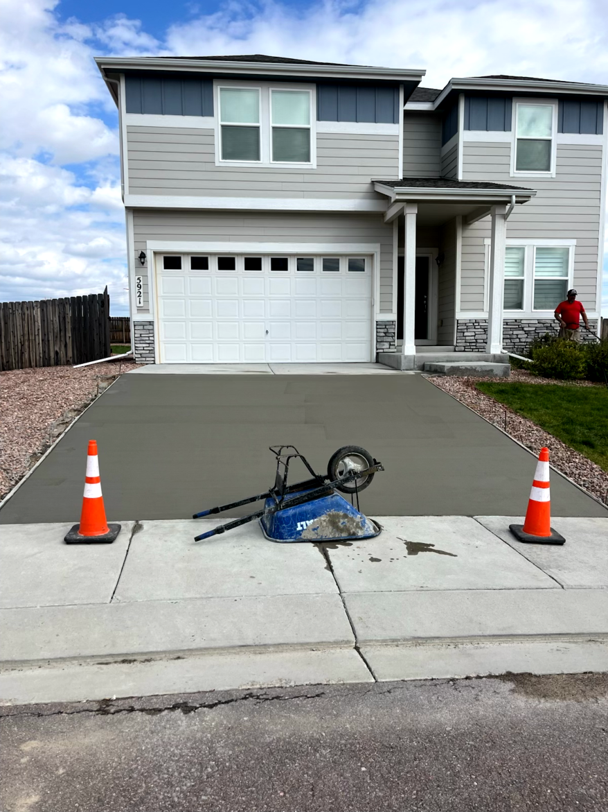 Newly poured concrete driveway in front of a two-story gray house. Construction cones and wheelbarrow present.