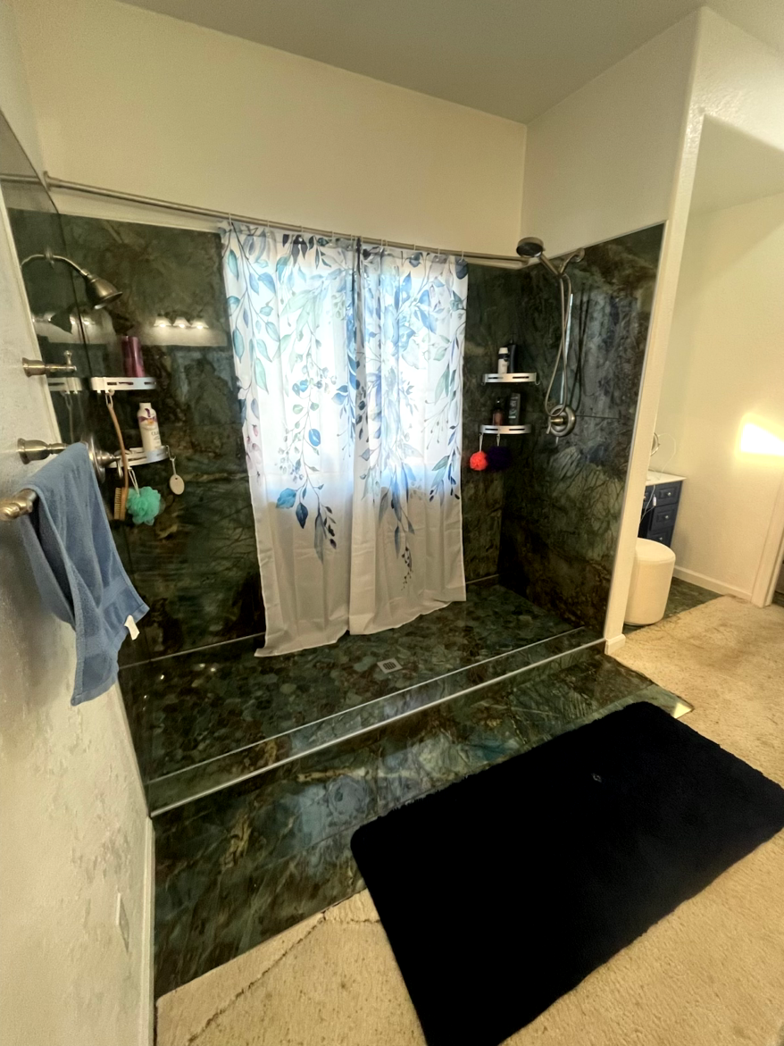 Green marble shower with floral shower curtain, shelves with toiletries, and a black bath mat.