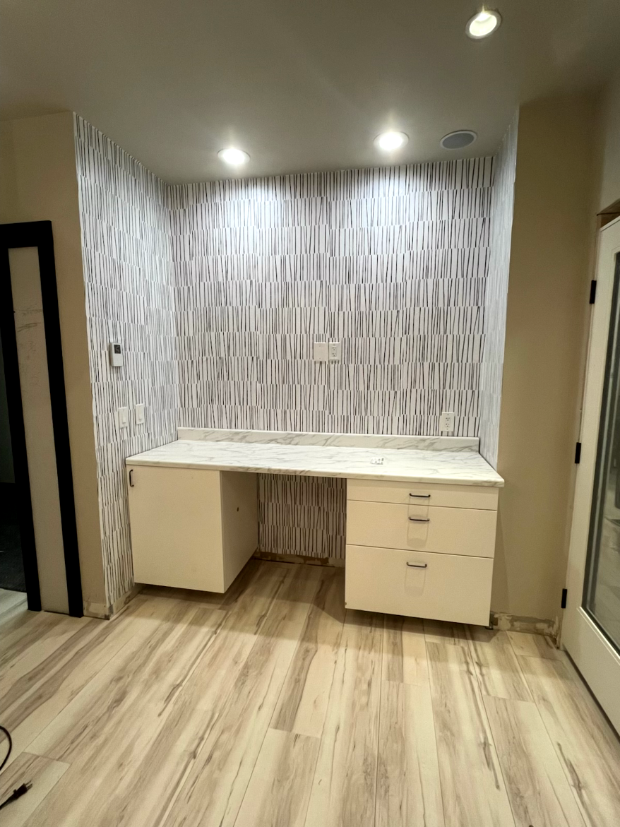 Built-in desk in alcove with patterned tile wall and recessed lighting. White cabinetry, light wood-look floor.
