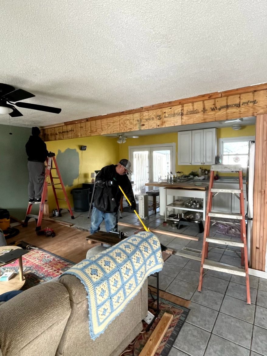 Two workers removing part of a ceiling beam in a home renovation. Interior shows a yellow wall, ladder, and debris.