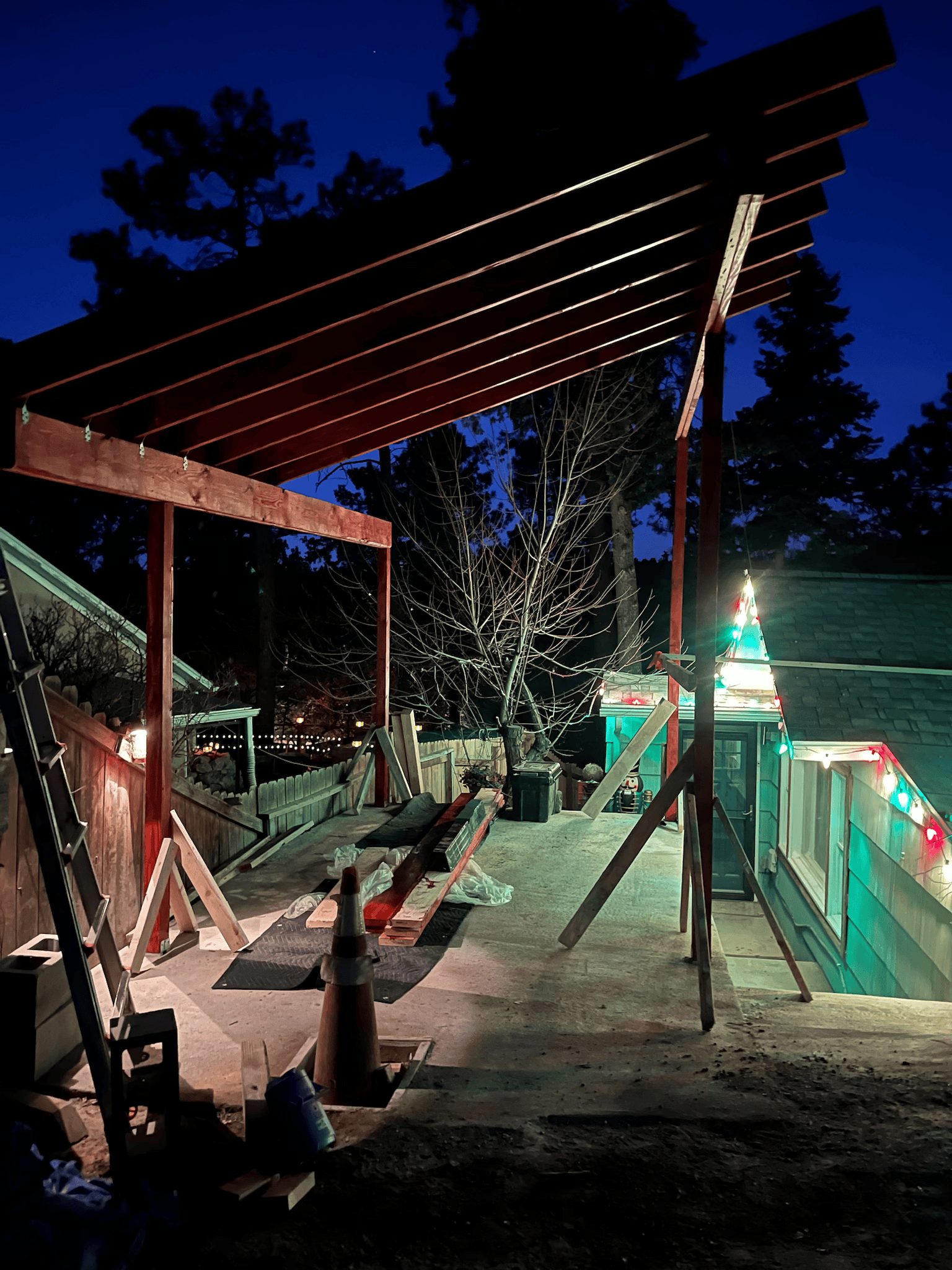 Wooden pergola under construction outdoors at dusk. Red-painted beams and stringers, against a dark blue sky.