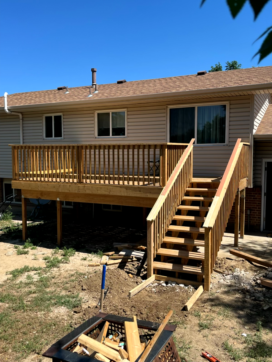Wooden deck with stairs attached to a beige house, blue sky, and a fire pit in the yard.