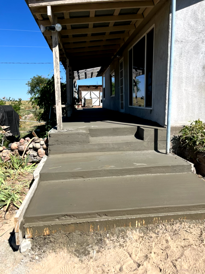 Concrete steps leading up to a house's covered porch, with a wooden trellis roof and a clear blue sky.