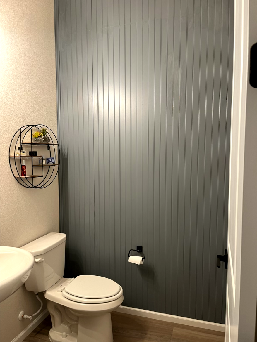 Bathroom with gray vertical panel wall, toilet, sink, and decorative shelf.