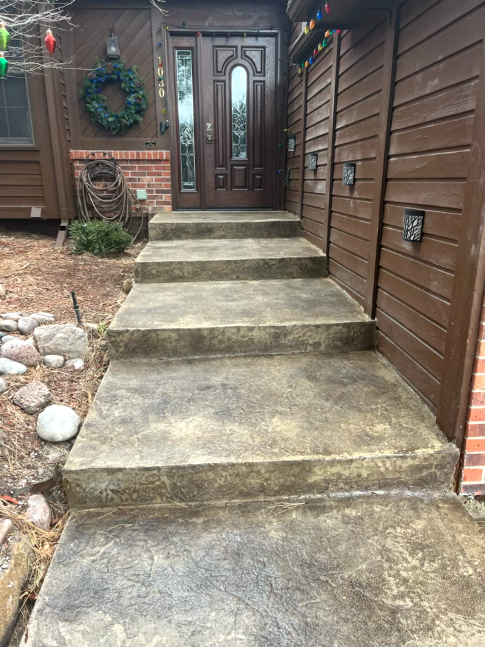 Concrete steps leading up to a brown front door. Wooden siding and a wreath are visible.