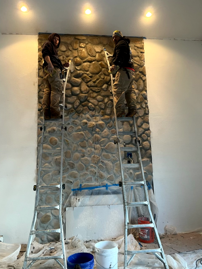 Two people on ladders working on a stone wall fireplace. Indoors, under bright lights.