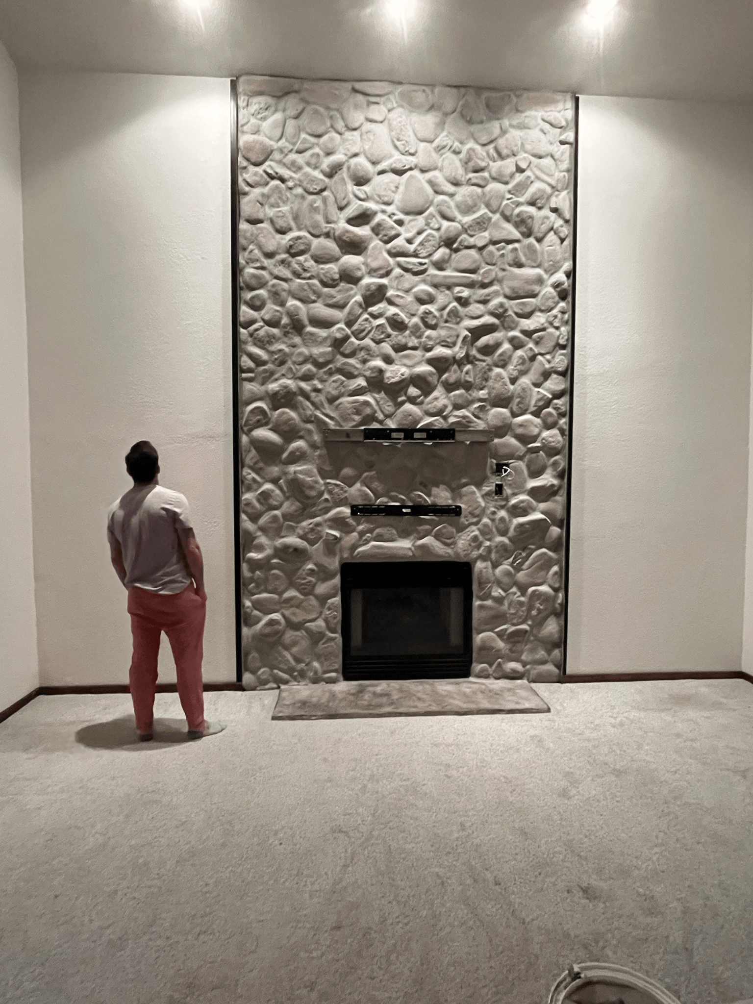 Person looking at a stone fireplace in an empty room with a light carpet.