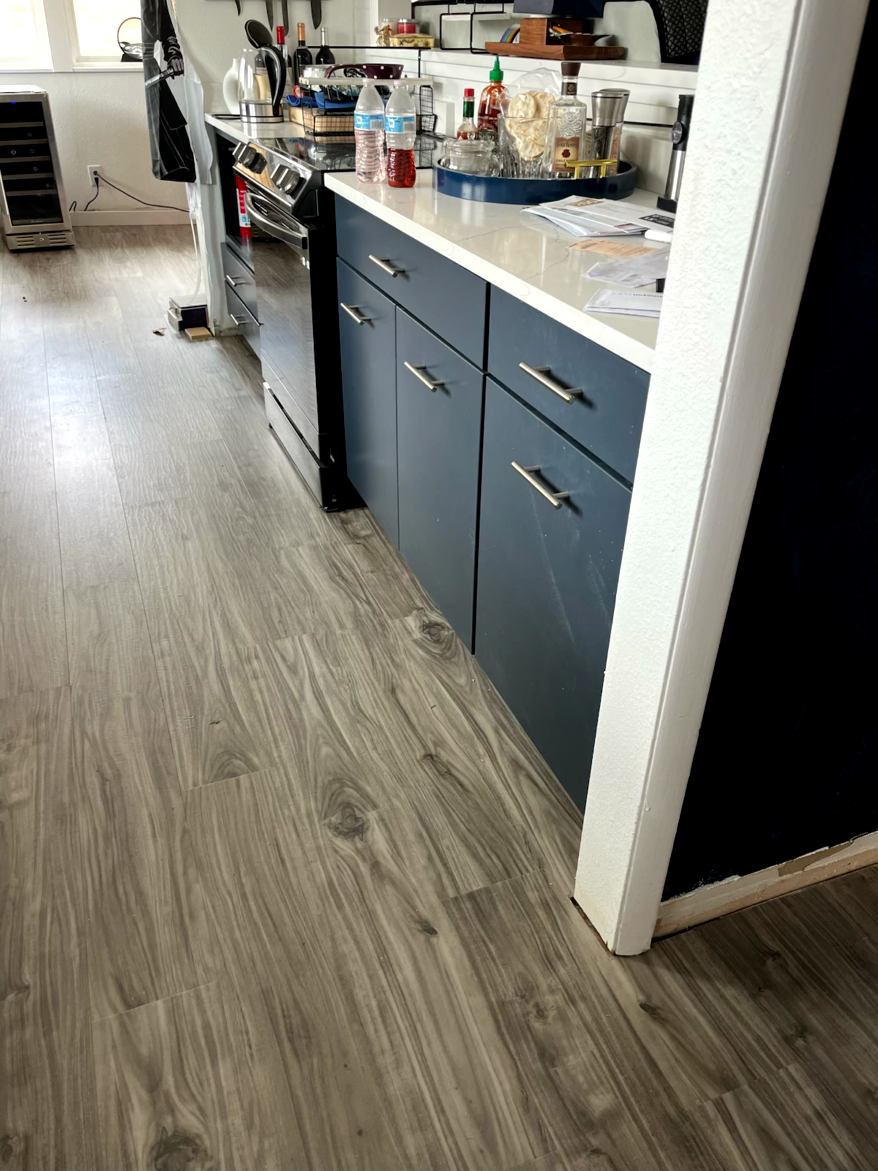 Kitchen with gray wood-look flooring, blue cabinets, white countertop. A stainless steel appliance and white wall are also visible.