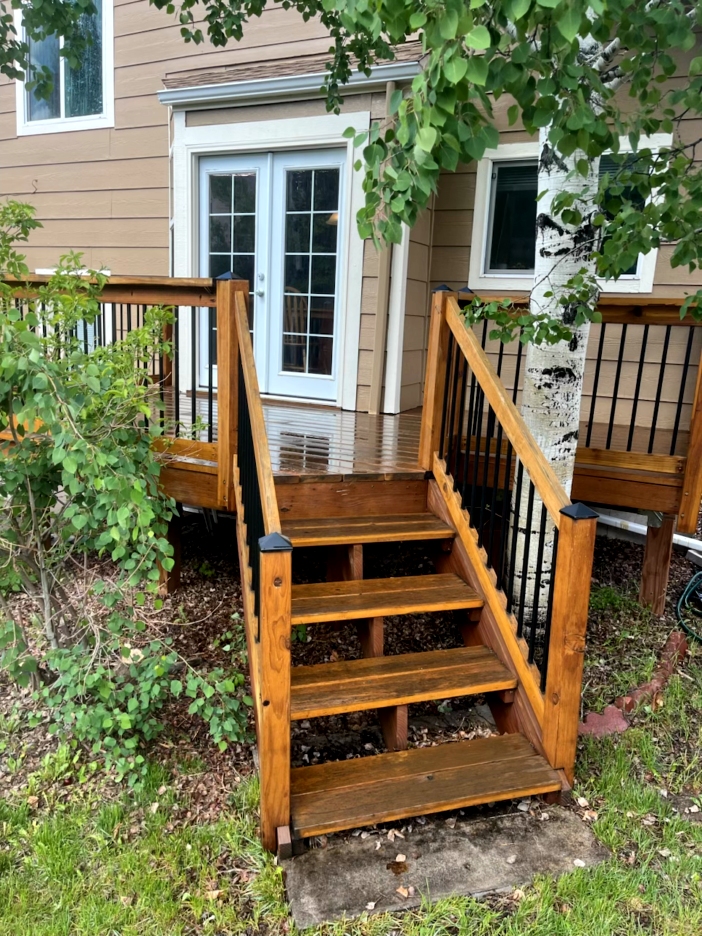 Wooden deck with stairs leading down to a lawn. Brown building with white double doors. Black railing.
