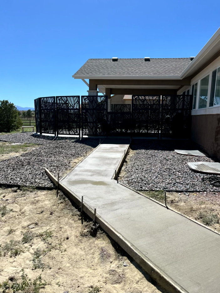 Concrete pathway leading to a deck with decorative black fencing. Gravel and landscaping surround it. Clear blue sky.