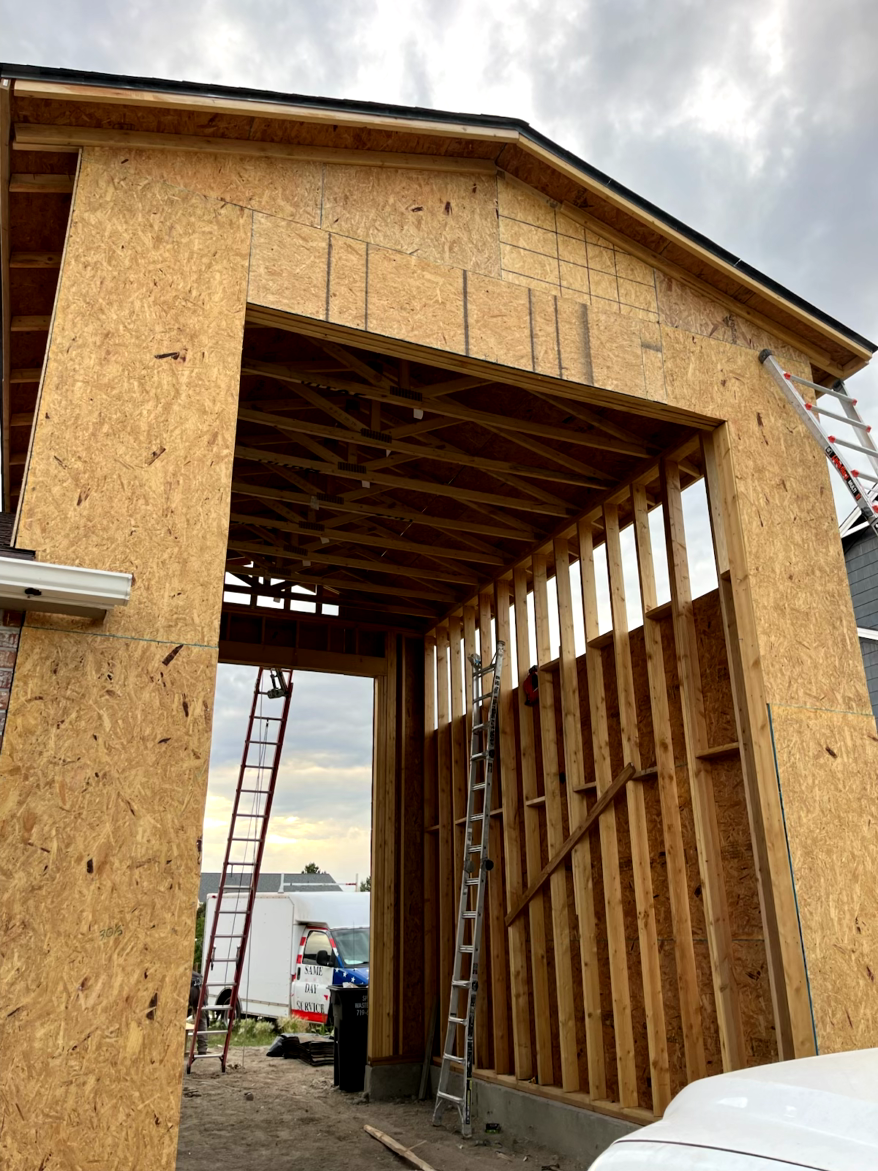 Garage under construction; wood framing and OSB sheathing visible, ladders, cloudy sky.