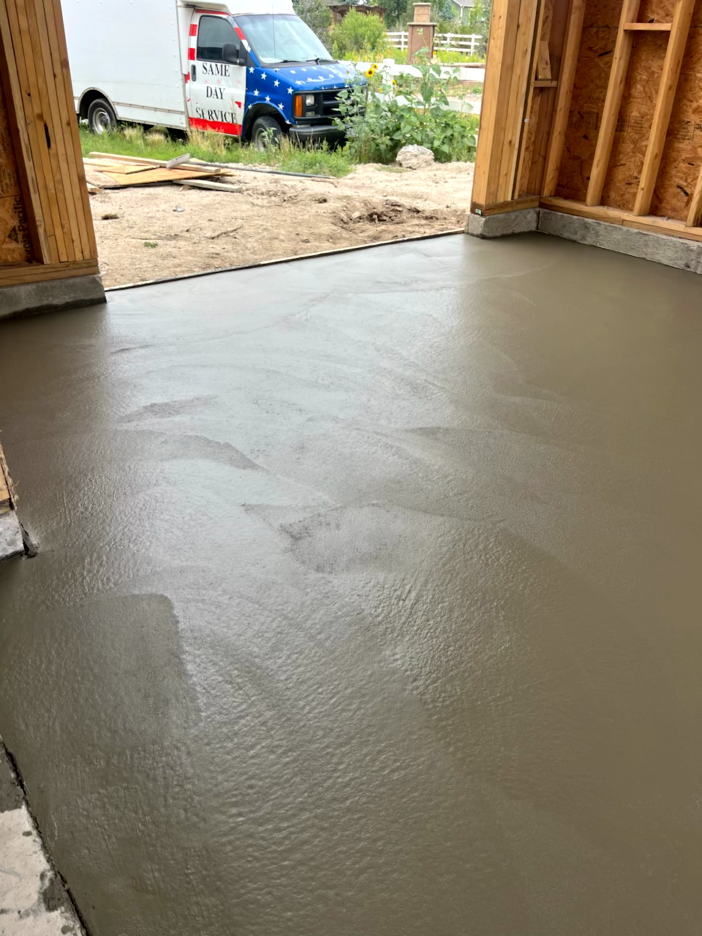 Newly poured gray concrete floor in a garage under construction. Wooden framing is visible. White truck with flag design in background.