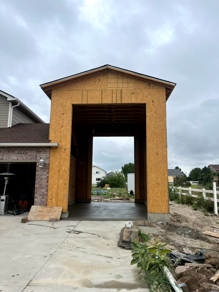 Framed garage under construction, open to the sky. Wooden beams, concrete floor, overcast day.