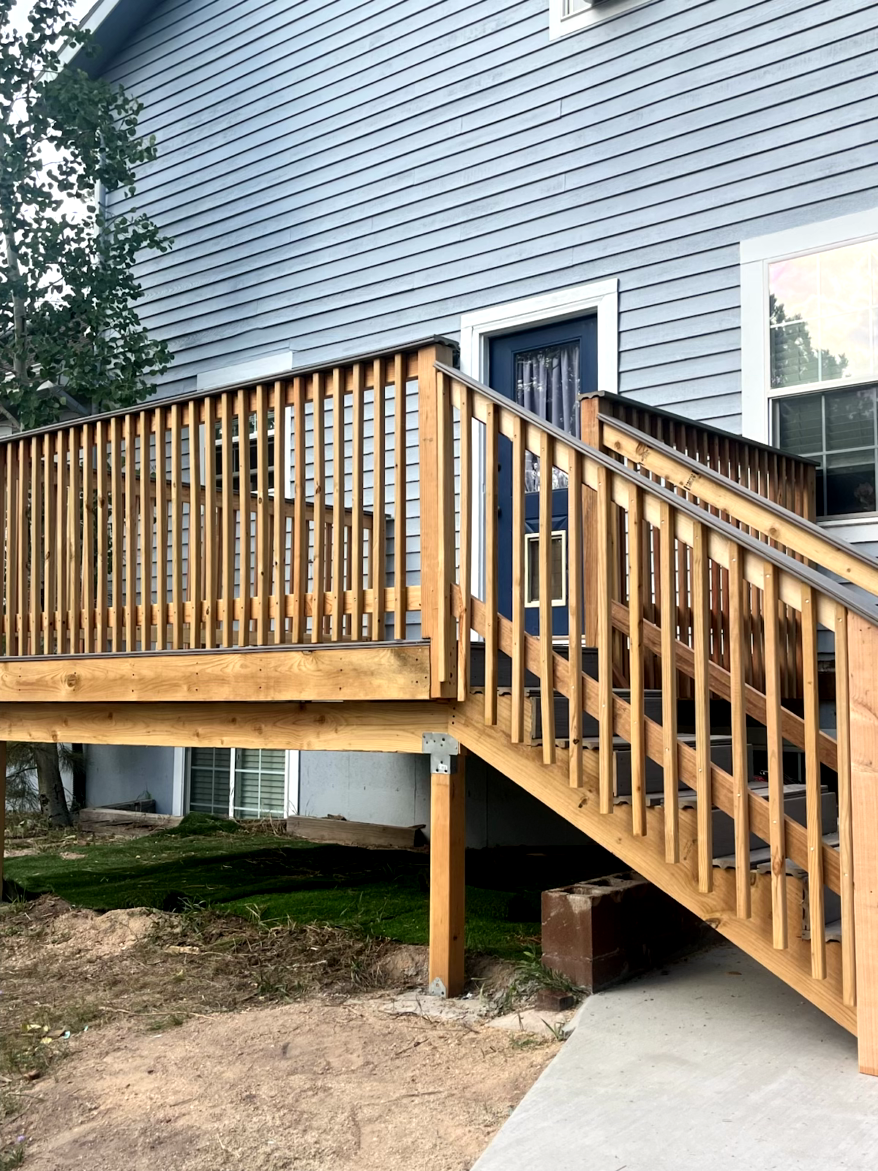 Wooden deck with stairs leading to a blue door on a gray-sided building. Brown railing and steps.