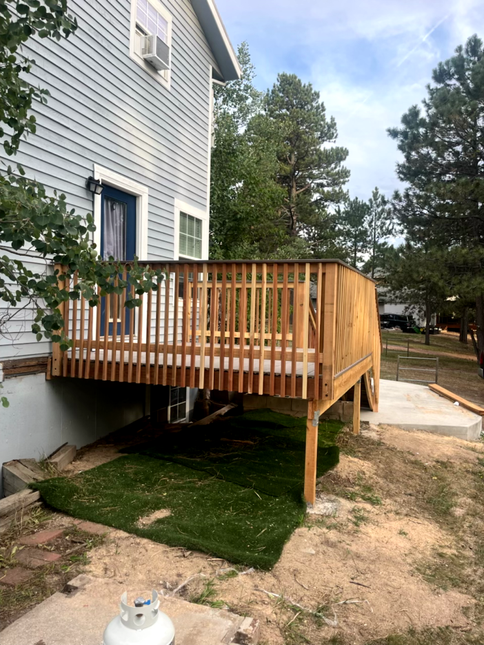 Wooden deck attached to a light blue house, overlooking a grassy area. A propane tank sits in the foreground.