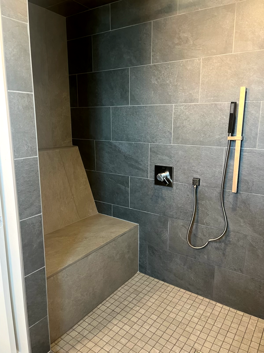 A modern shower with dark gray tiled walls and a built-in seat. A handheld showerhead is mounted on the wall.