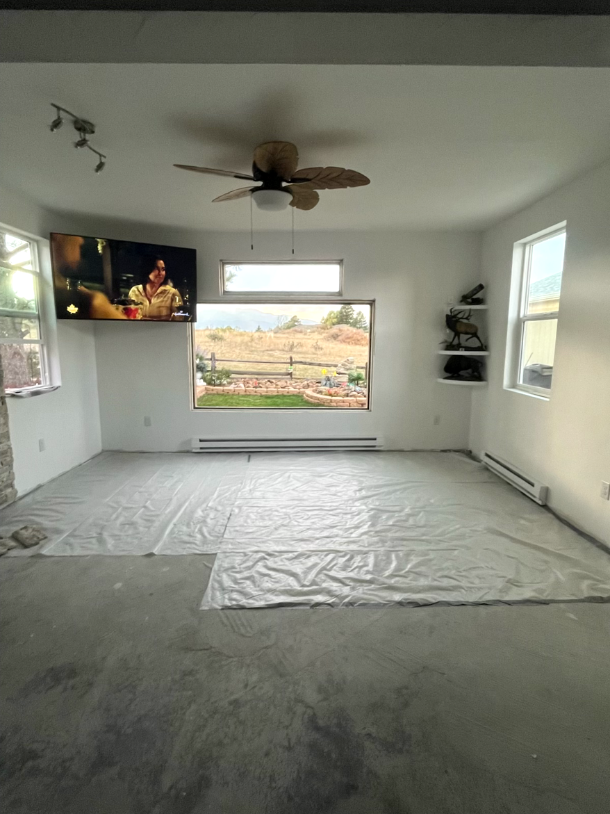 Empty white living room with concrete floor, large window, TV, and ceiling fan.