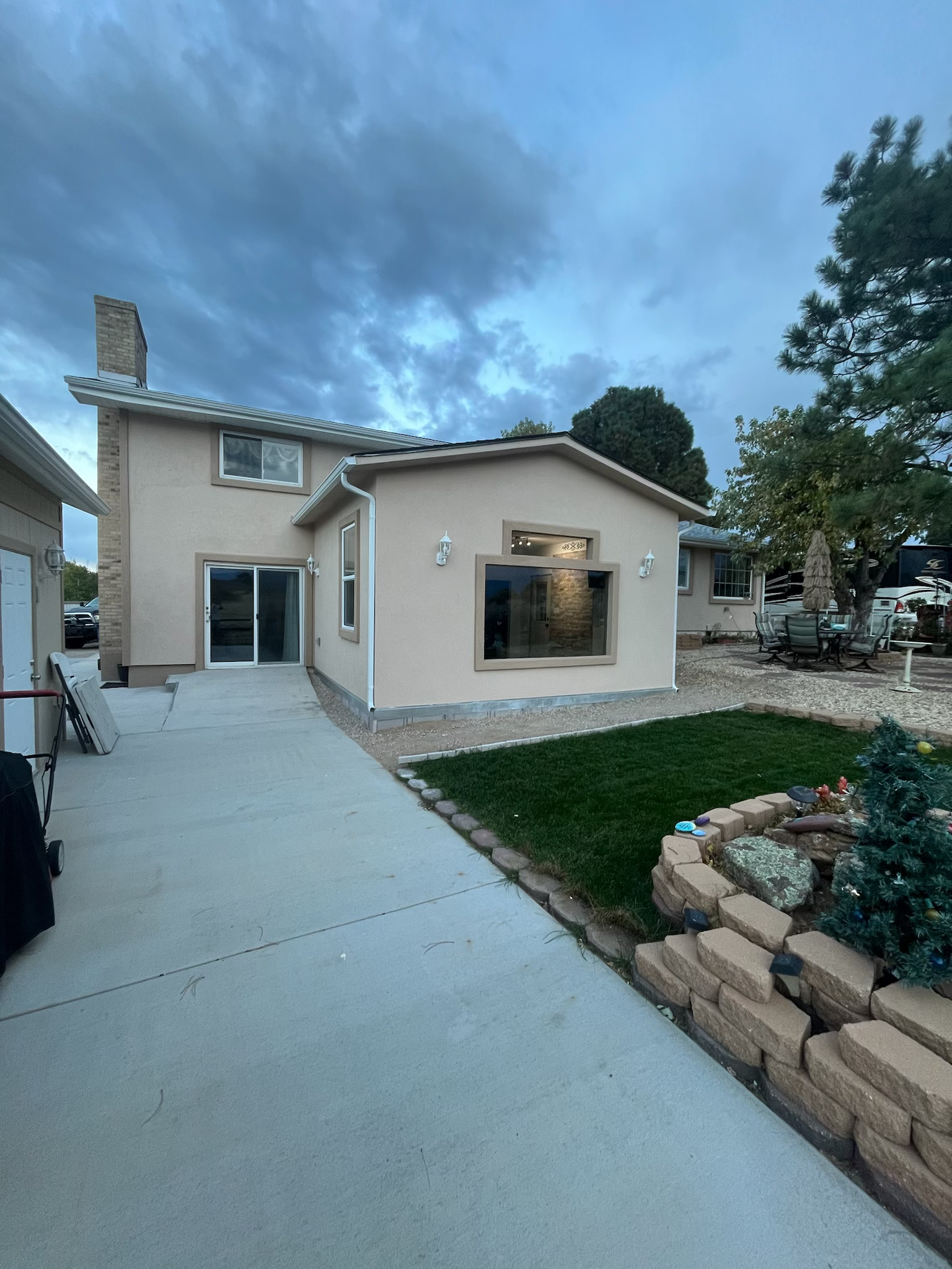 Backyard of a tan house with a concrete path, small lawn, and a large window; cloudy sky.