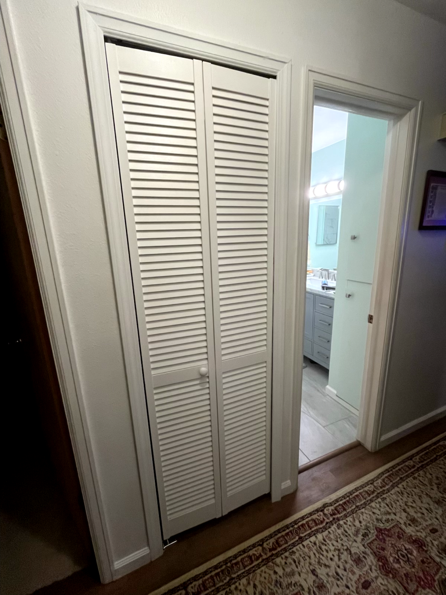 White louvered closet doors next to a doorway leading to a light blue bathroom. Brown floor with a rug.