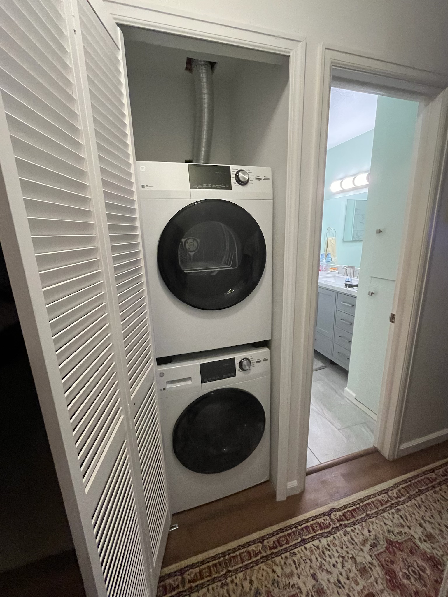 Stacked white washer and dryer in a closet, next to a bathroom doorway. Slatted white doors are open.