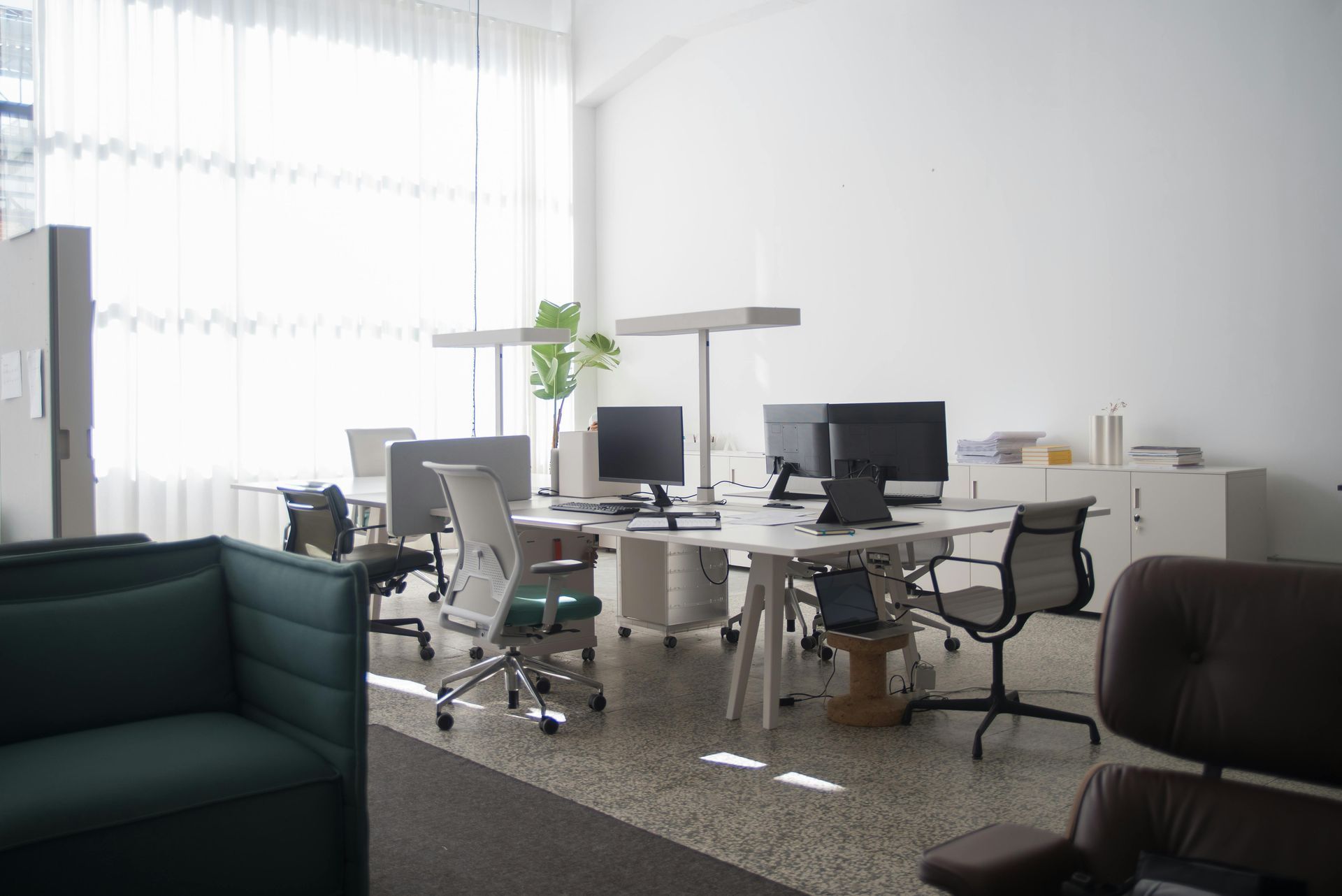 Modern office space with desks, chairs, computers, and a sofa near a large window, bathed in sunlight.
