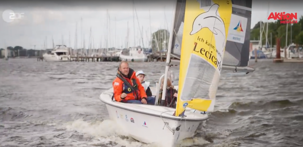 Kleines Segelboot mit einem orange gekleideten Segler rast auf unruhigem Wasser in der Nähe eines Yachthafens.
