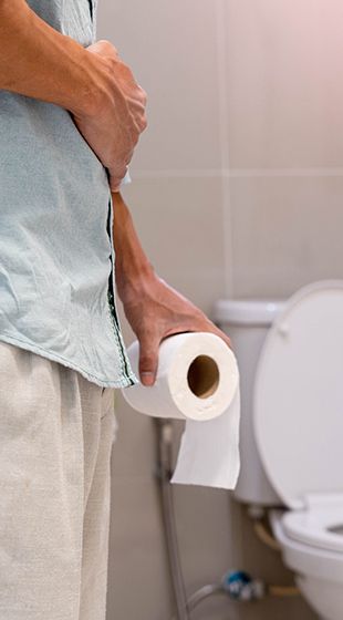 Mann hält sich den Bauch und hält Toilettenpapier in der Nähe einer offenen Toilette in einem Badezimmer.