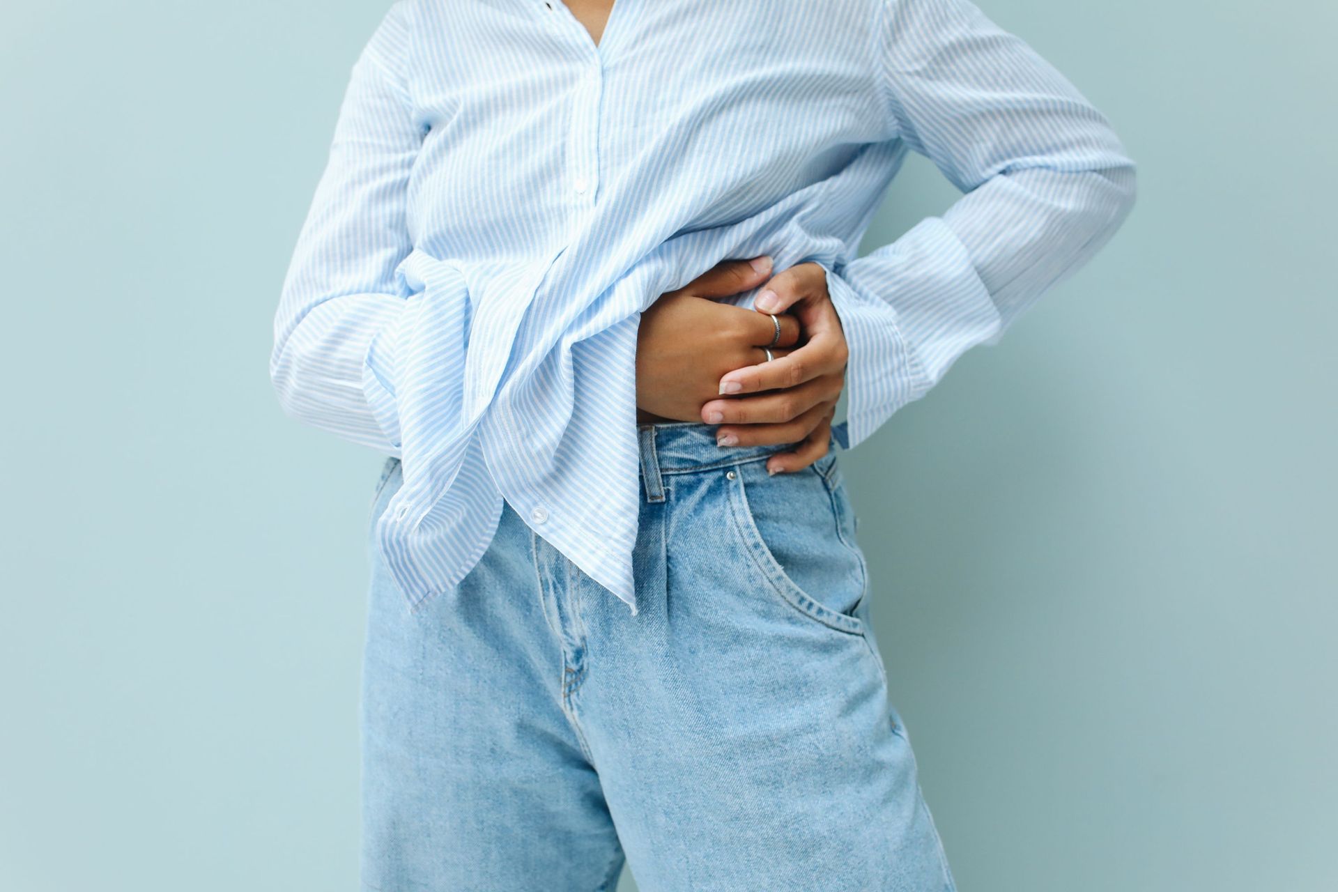 Eine Person hält sich in einem blau-weiß gestreiften Hemd und hellen Jeans den Unterbauch, was auf Magenschmerzen hindeutet.