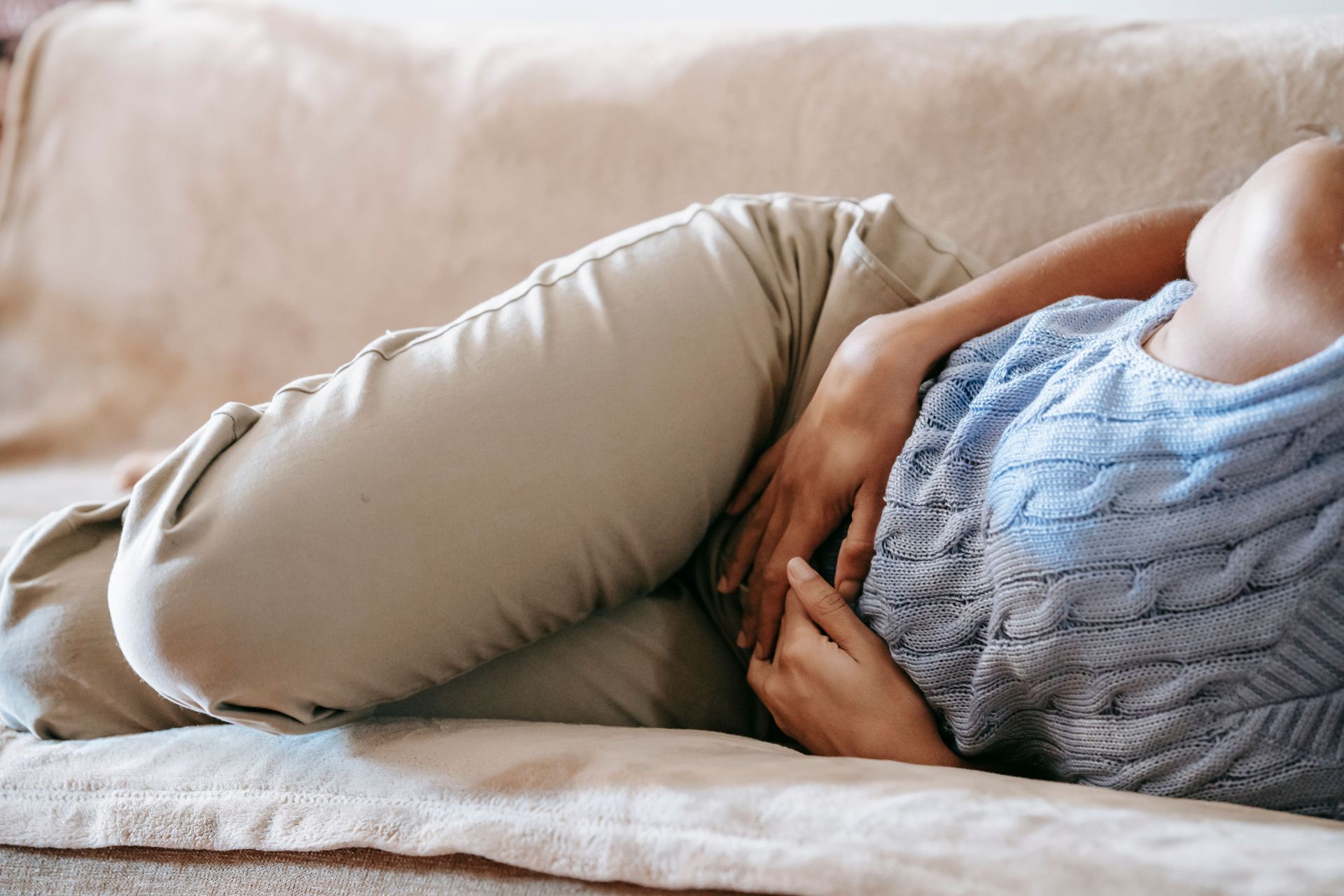 Frau mit weißer Hose und blauem Top liegt auf der Couch und hält sich den Bauch.