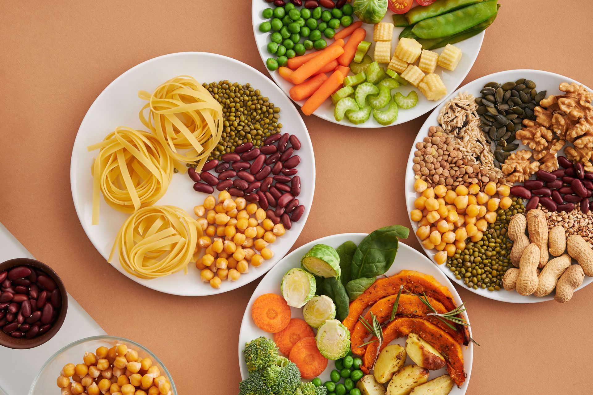 Assorted colorful vegetables, beans, grains, and pasta arranged on white plates on a peach background