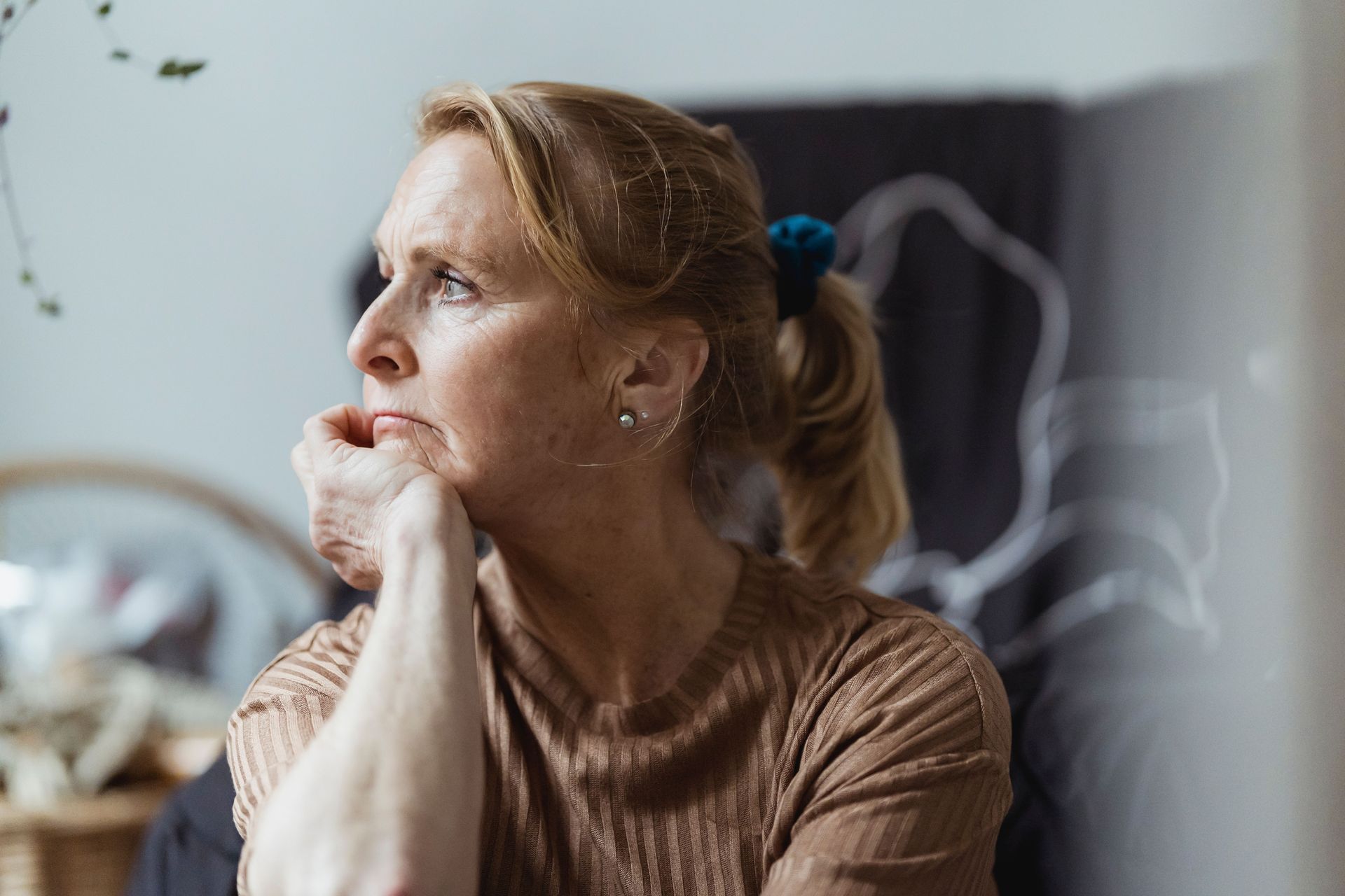 Woman in a brown sweater gazes thoughtfully out a window, resting her chin on her hands.
