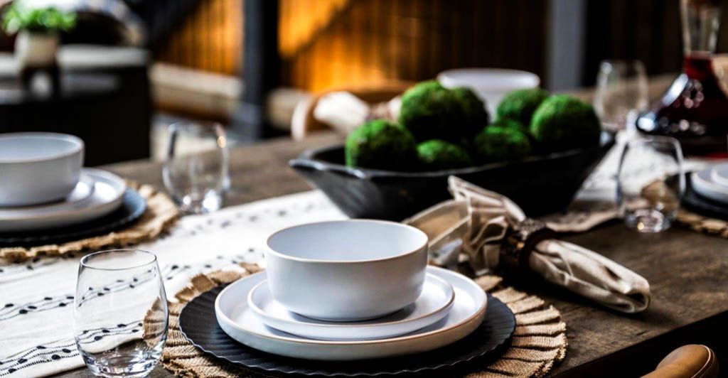 A table set for a dinner party with plates , bowls , glasses and a bowl of moss balls to present for a photo.