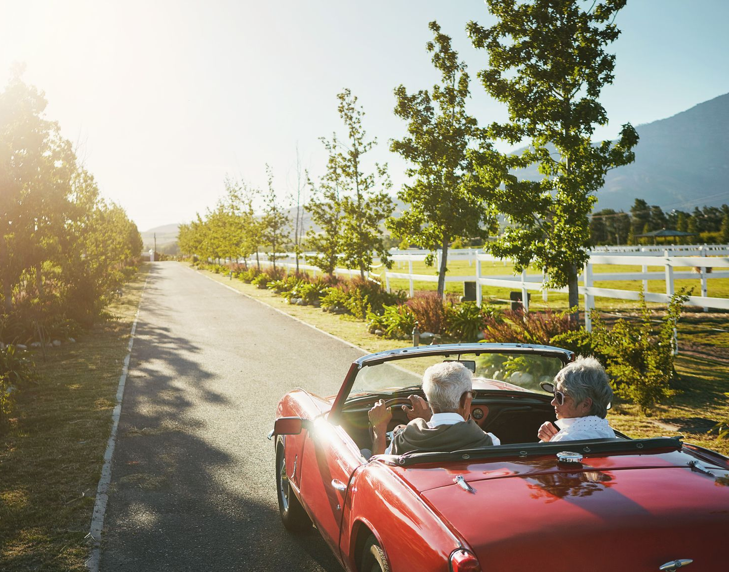 Two people in a red convertible driving down a sunny road lined with trees.
