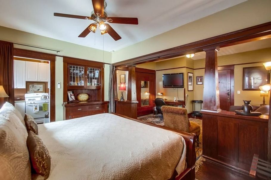 A cozy bedroom with a large, made bed in the foreground, wooden architectural details, and a view into a living area.