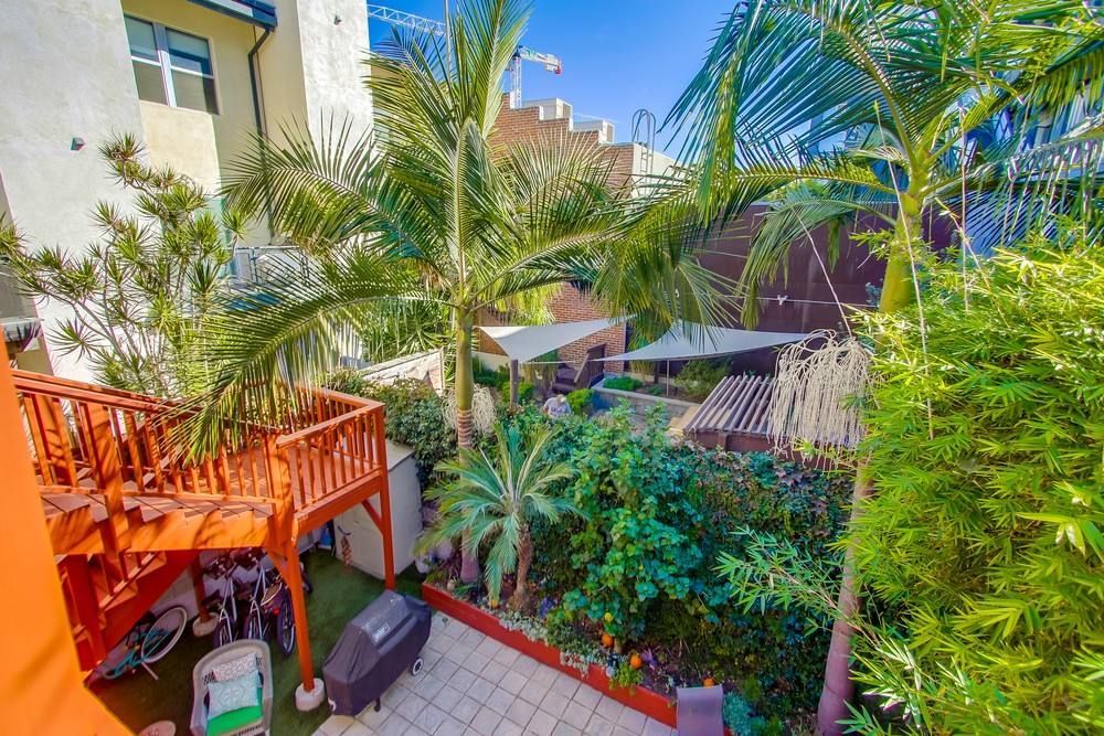 A wooden deck overlooking a lush courtyard with palm trees, green plants, a stone patio, and sail shades.