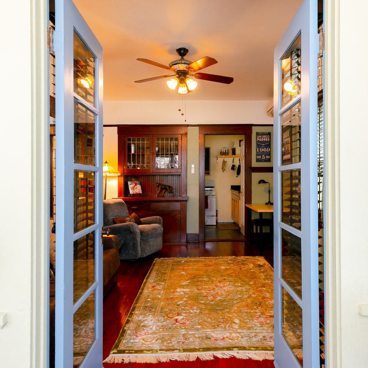 A view through blue-framed French doors into a cozy room with a patterned area rug, a plush armchair, and a ceiling fan.