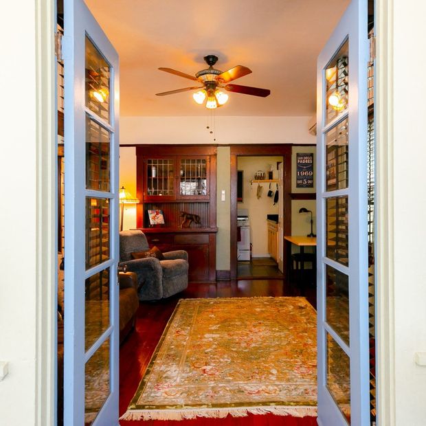 A view through light blue French doors into a living room with a large patterned rug, wood furniture, and a ceiling fan.