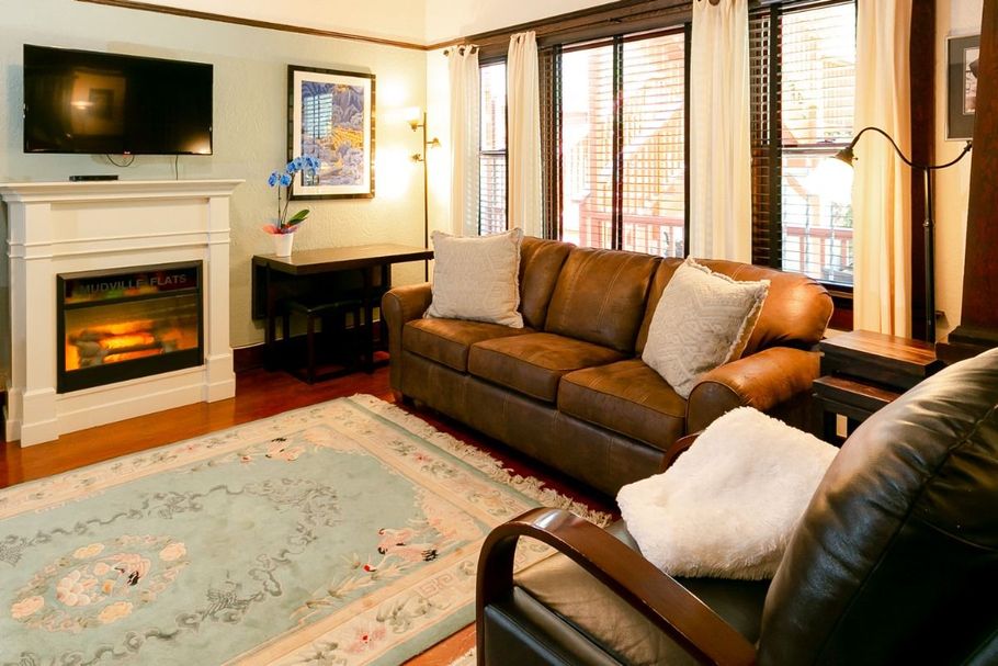 A cozy living room with a brown leather sofa, an electric fireplace, a wall-mounted TV, and a light blue patterned rug.