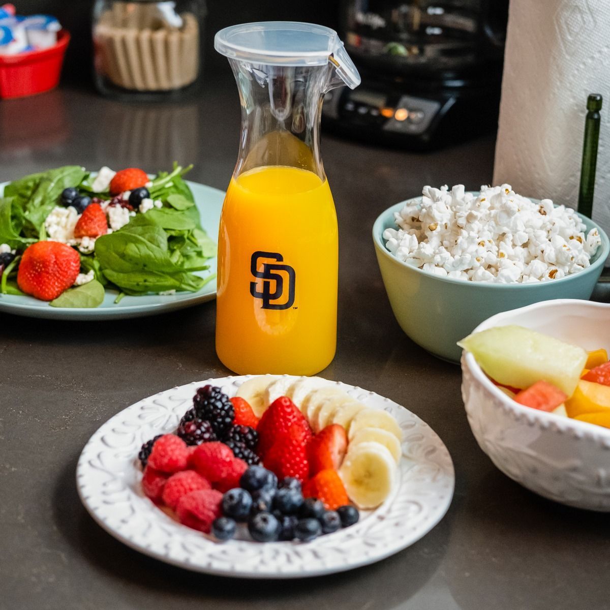 A healthy meal setup featuring a salad, a bowl of popcorn, a fruit plate with berries and banana, and orange juice.