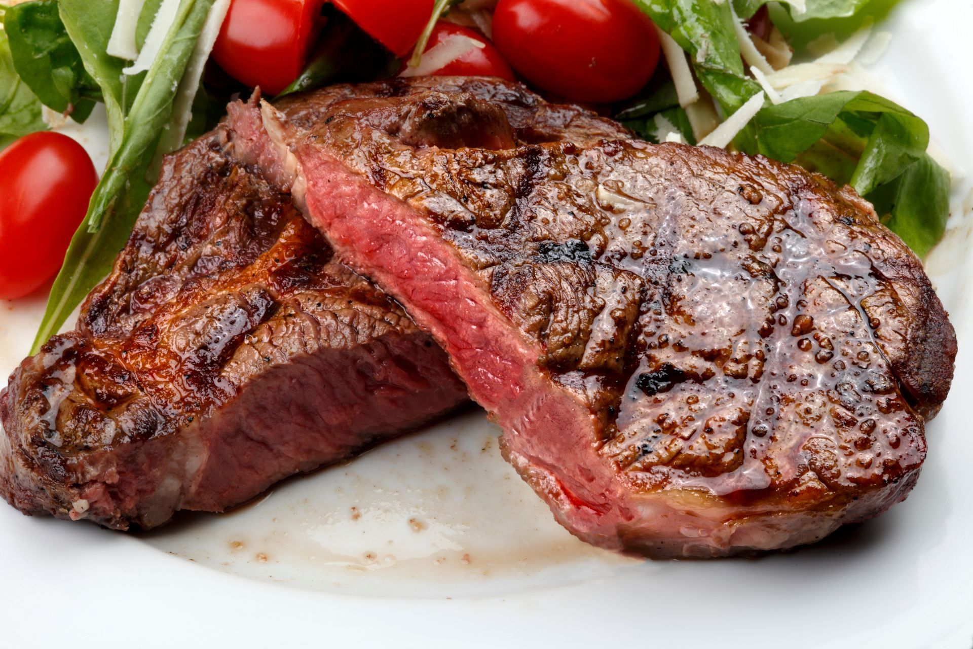 Grilled steak sliced to reveal a medium-rare pink center, served on a white plate with fresh salad and cherry tomatoes.