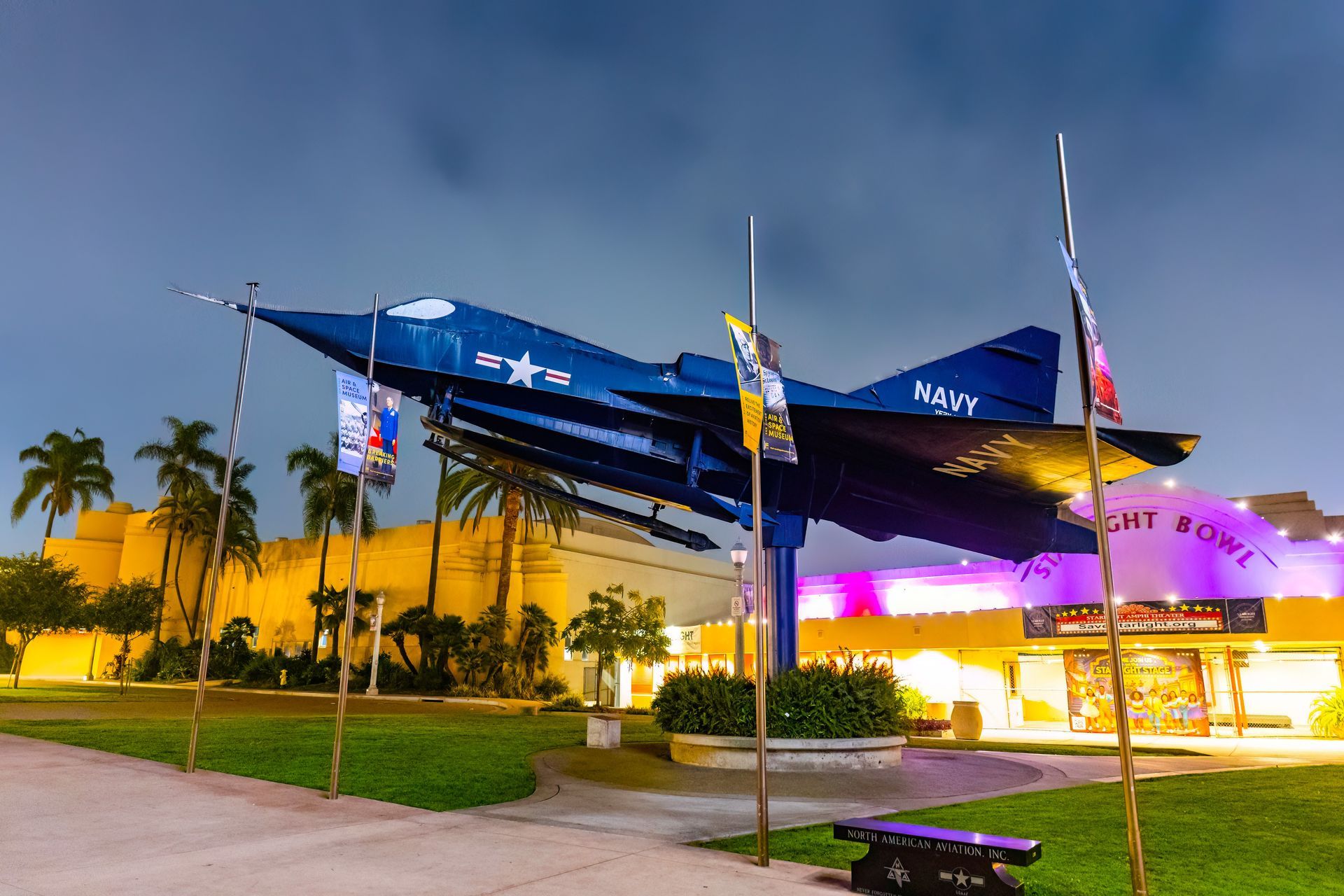 A dark blue fighter jet monument stands prominently on a pedestal at night in front of a brightly lit building.