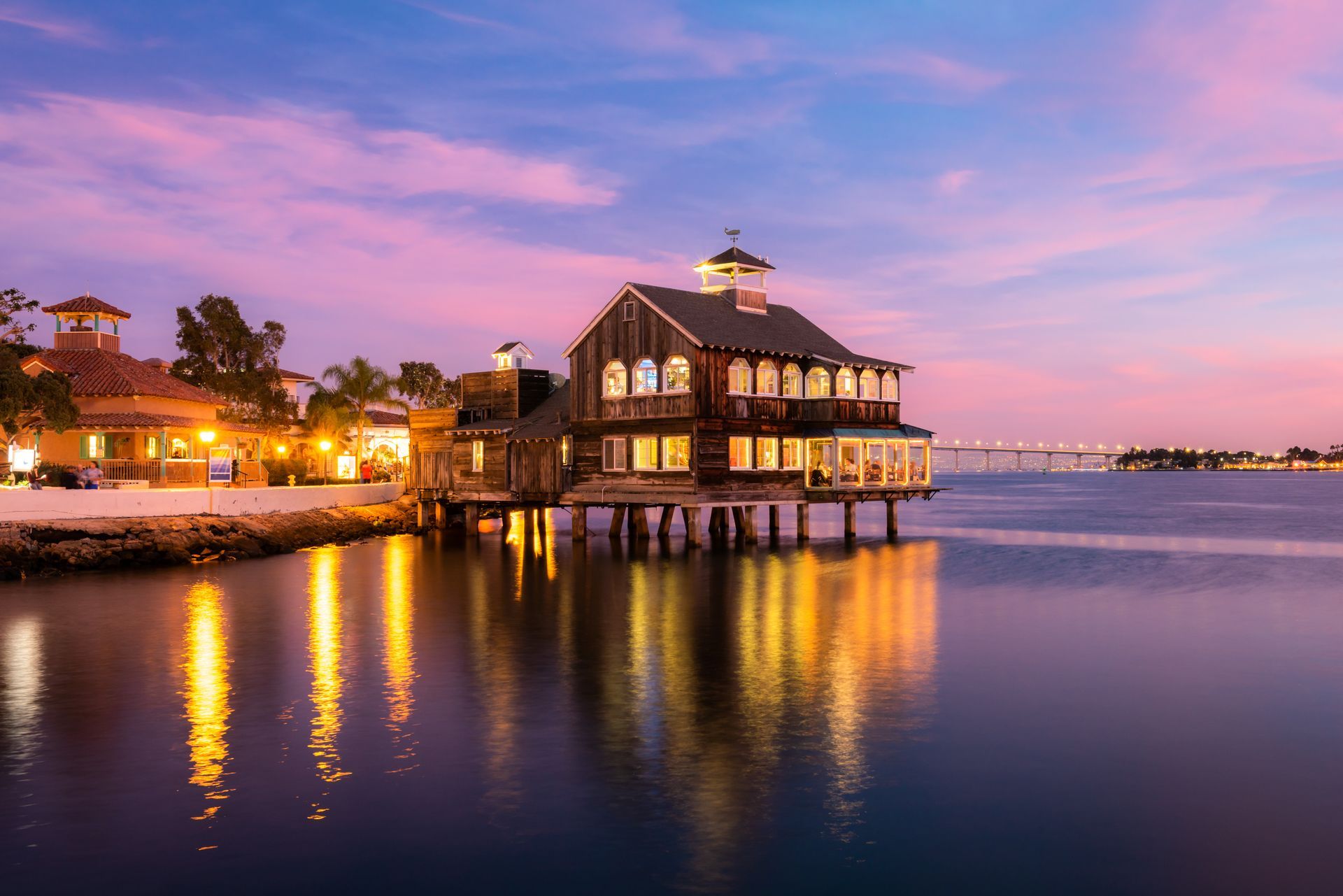 A stilted wooden restaurant building glows over calm bay water at dusk, under a vibrant pink and purple sunset sky.