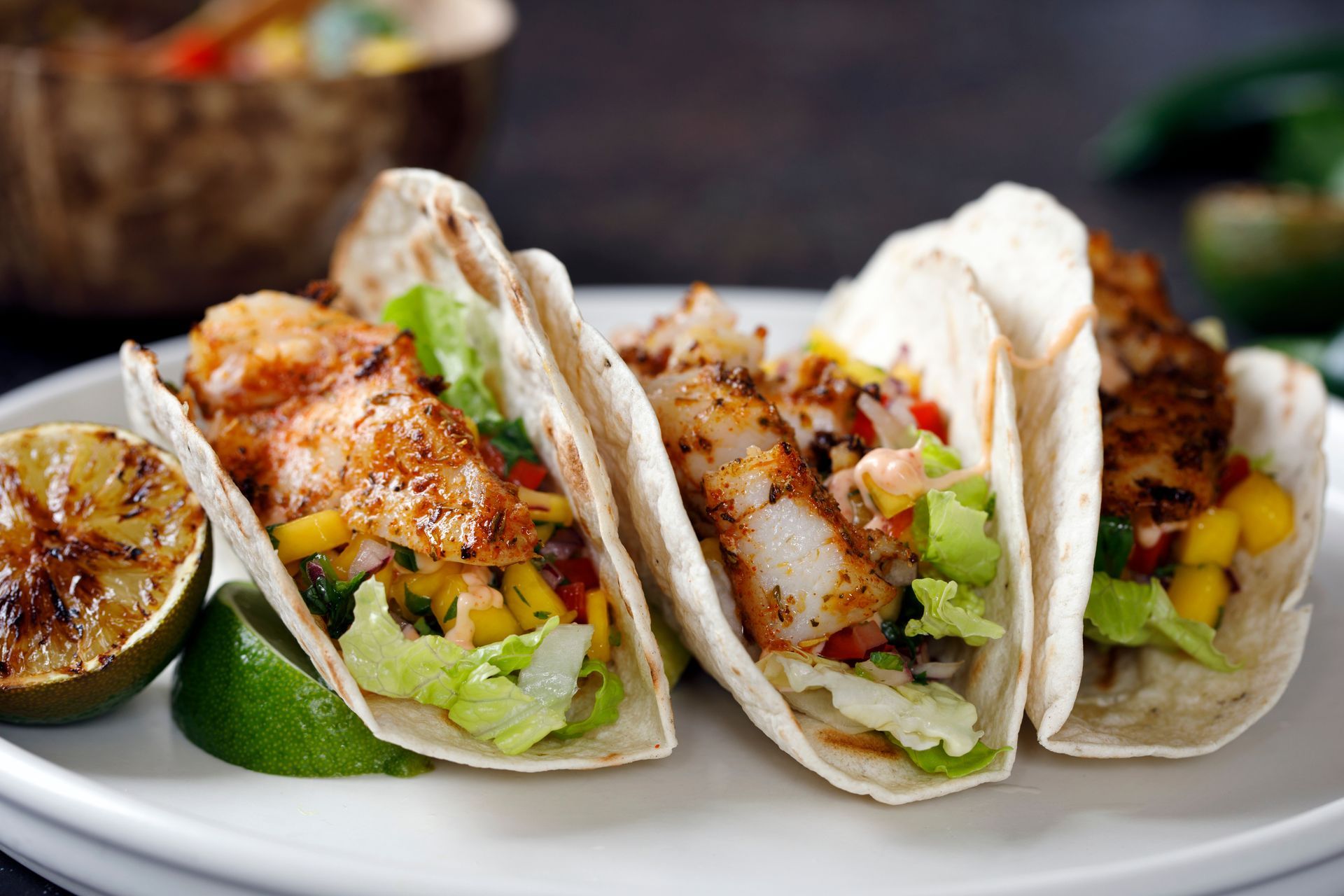 Three fish tacos on a white plate with a side of lime wedges, garnished with fresh salsa and shredded lettuce.